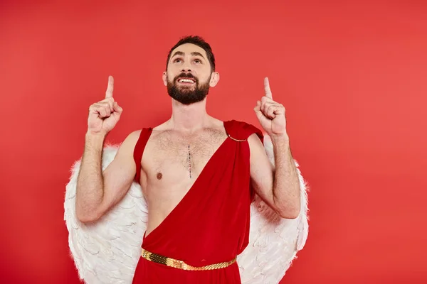 Fröhlicher bärtiger Amor, der nach oben schaut und mit den Fingern auf die rote Kostümparty der Valentins zeigt — Stockfoto