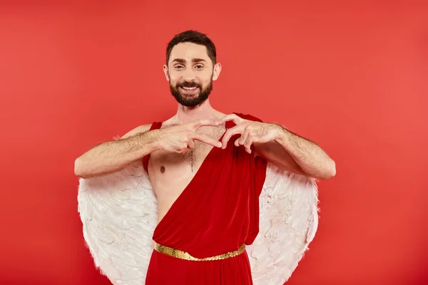 Erfreut bärtiger Amor Mann zeigt Herzzeichen mit Fingern und lächelt in die Kamera auf rotem Hintergrund — Stockfoto