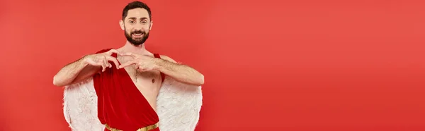 Homem barbudo alegre se veste como cupido mostrando sinal de coração e olhando para a câmera no vermelho, banner — Fotografia de Stock