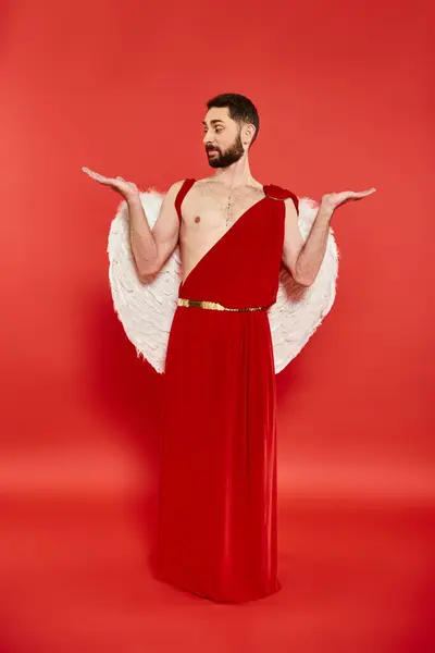Comprimento total do homem vestido como cupido olhando para suas palmas abertas no vermelho, dia de São Valentim — Fotografia de Stock