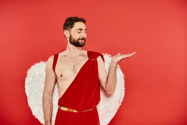 Smiling bearded cupid man pointing with hand with open palm on red, st valentines costume party — Stock Photo