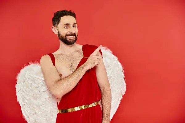 Cheerful bearded cupid man looking at camera and pointing away with finger on red, st valentines day — Stock Photo