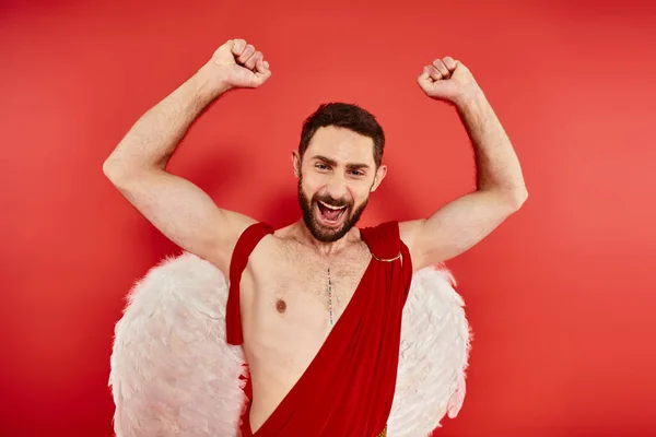 Excited bearded cupid man rejoicing and showing win gesture with raised hands on red backdrop — Stock Photo
