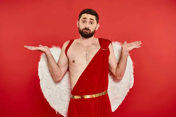 Confused bearded man in cupid costume showing shrug gesture and looking at camera on red backdrop — Stock Photo