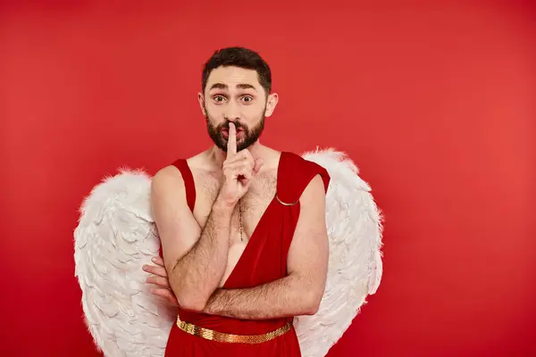 Excité homme barbu en costume de Cupidon montrant le signe shh et regardant la caméra sur rouge, st valentines — Photo de stock