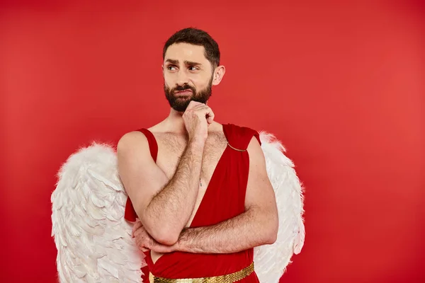 Displeased bearded man in cupid costume holding hand near chin and  looking away on red backdrop — Stock Photo