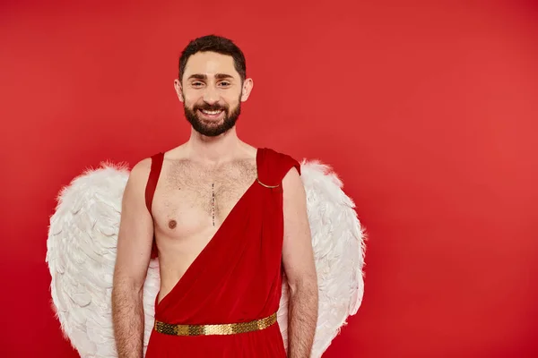 Hombre barbudo positivo vestido como Cupido con alas sonriendo a la cámara en rojo, San Valentín día - foto de stock