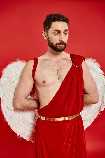 Pensive bearded man dressed as cupid with wings standing with hands behind back on red backdrop — Stock Photo