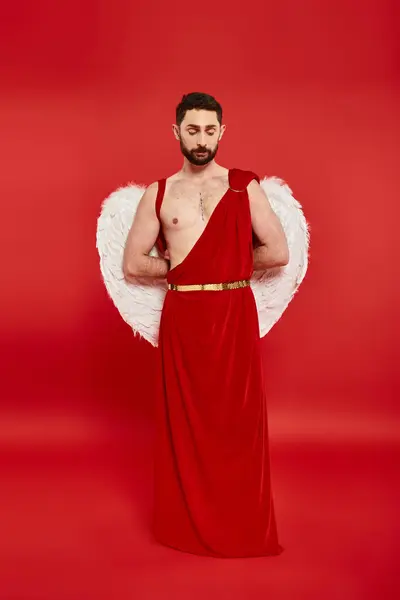 Full length of thoughtful bearded man in cupid costume standing with hands behind back on red — Stock Photo