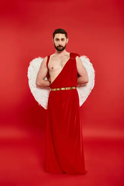 Longitud completa del hombre barbudo en traje de Cupido de pie con las manos detrás de la espalda mirando a la cámara en rojo - foto de stock
