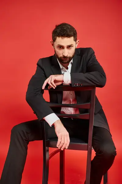 Серйозний гострий одягнений чоловік сидить на стільці і дивиться на камеру на червоному, успішний бізнесмен — стокове фото