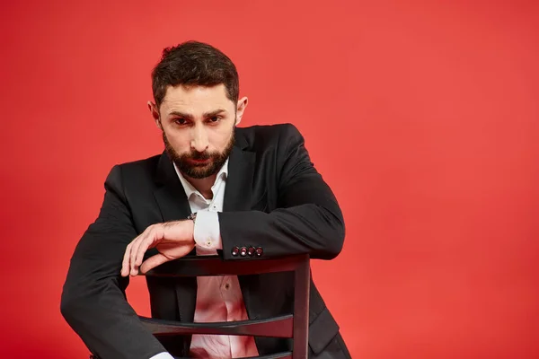 Вдумливий бородатий бізнесмен в чорному костюмі сидить на стільці на червоному тлі, корпоративний лідер — стокове фото