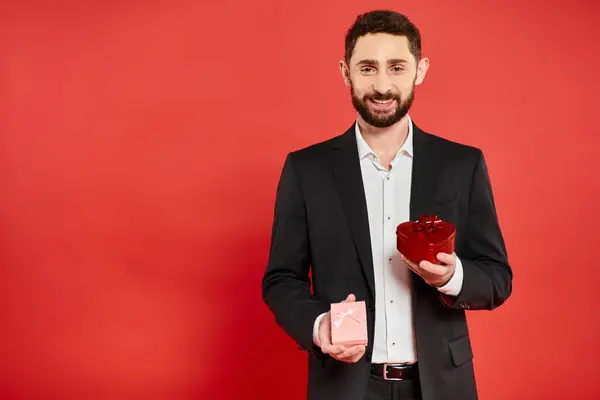 Alegre hombre de negocios barbudo en traje elegante negro sosteniendo cajas de regalo en rojo, regalos de San Valentín - foto de stock