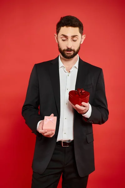 Homme barbu réfléchi en noir tenue formelle en choisissant Saint-Valentin présent sur fond rouge — Photo de stock