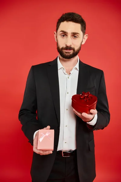 Hombre barbudo positivo en traje elegante celebración de San Valentín día regalos y mirando a la cámara en rojo - foto de stock