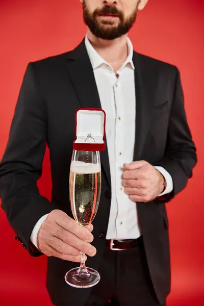 Vue recadrée de l'homme en costume noir avec verre de champagne et boîte cadeau avec bague à bijoux sur rouge — Photo de stock
