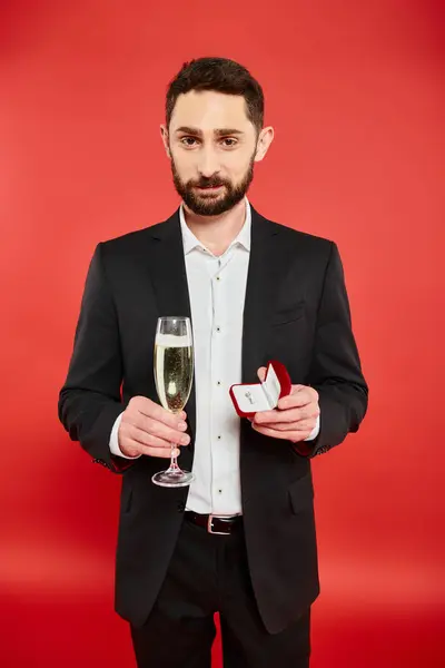 Усміхнений бородатий чоловік в елегантному вбранні з шампанським та ювелірною коробкою на червоному, день Святого Валентина — стокове фото