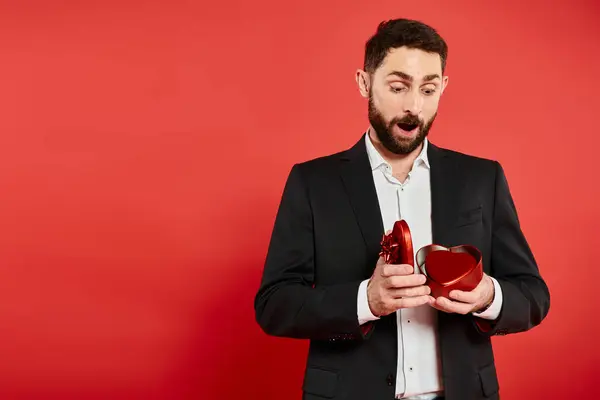 Amazed man in black suit opening heart-shaped gift box on red, Saint Valentines day present — Stock Photo