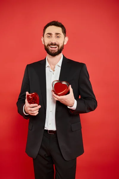 Excité homme barbu en costume noir avec boîte cadeau en forme de coeur rire sur le rouge, st Saint-Valentin — Photo de stock