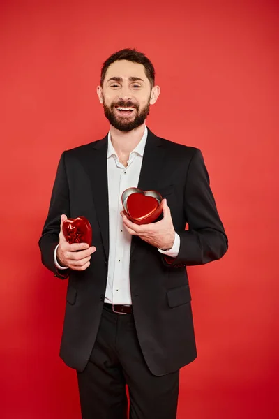 Excited bearded man in black suit with heart-shaped gift box laughing on red, st valentines day — Stock Photo