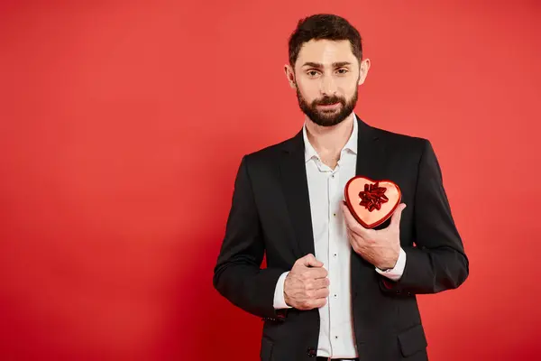 Pleased bearded businessman in black suit with heart-shaped gift box on red, Saint Valentines day — Stock Photo