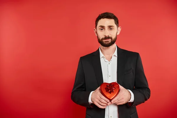 Homem barbudo vestido afiado segurando Dia dos Namorados st presente na caixa de presente em forma de coração no vermelho — Fotografia de Stock