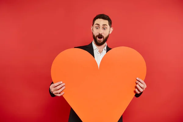 Homem barbudo espantado com boca aberta e enorme coração de papel laranja no vermelho, conceito de São Valentim — Fotografia de Stock