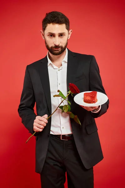 Confiado hombre elegante con rosa y delicioso pastel en forma de corazón en rojo, celebración de San Valentín - foto de stock
