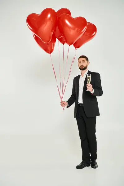 Uomo ricco in abito nero con champagne e palloncini rossi a forma di cuore su grigio, giorno st valentines — Foto stock