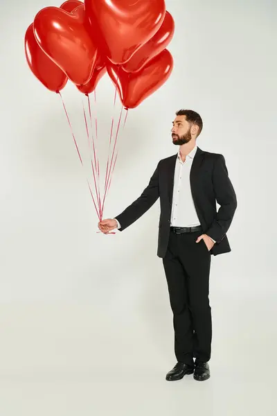 Intera lunghezza dell'uomo in abito nero elegante con palloncini rossi a forma di cuore e mano in tasca su grigio — Foto stock