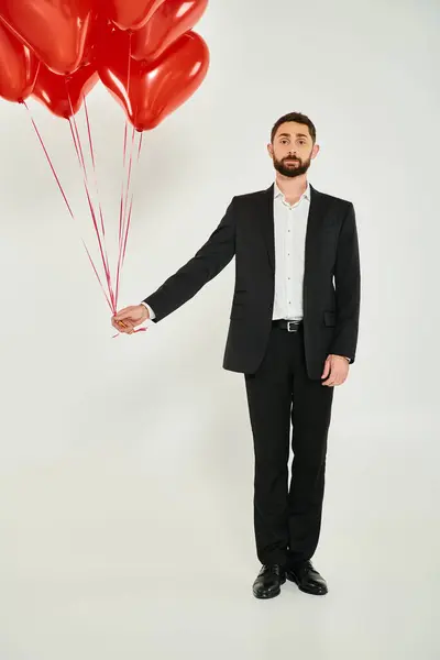Hombre de negocios barbudo en traje negro con globos rojos en forma de corazón mirando a la cámara en gris - foto de stock