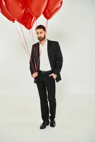 Bell'uomo barbuto in completo nero con palloncini rossi a forma di cuore e mano in tasca su grigio — Foto stock