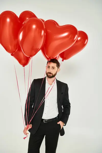 Bearded man in formal wear with heart-shaped balloons and hand in pocket on grey, st valentines day — Stock Photo