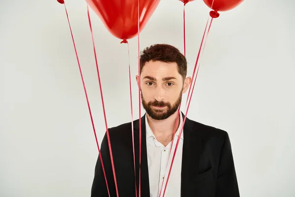 Charismatique homme barbu regardant la caméra près de ballons festifs rouges sur gris, concept st valentines — Photo de stock