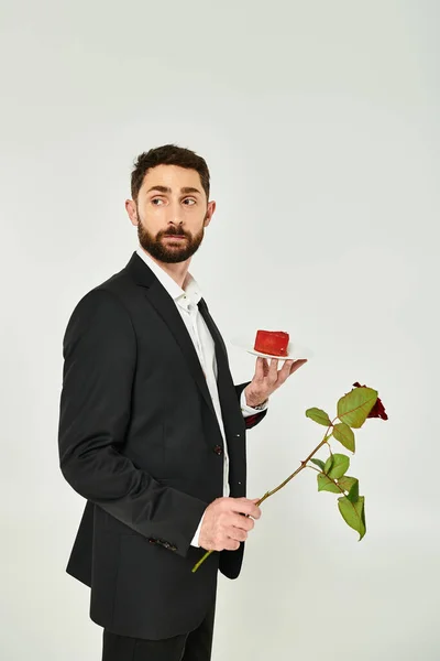 Elegante uomo barbuto con torta a forma di cuore e rosa rossa che distoglie lo sguardo sulla vacanza grigia, st San Valentino — Foto stock