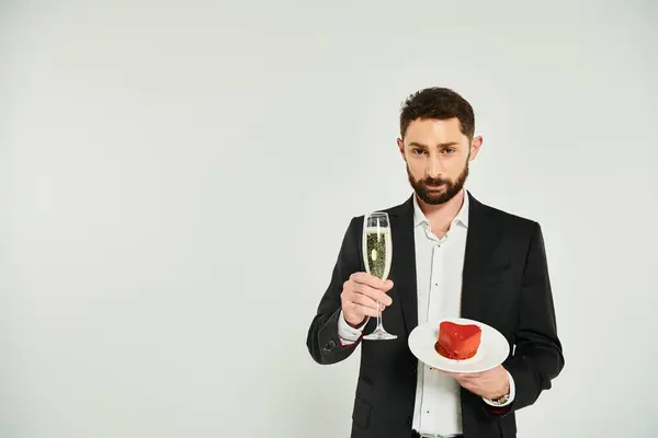 Красивый элегантный мужчина с бокалом шампанского и вкусным пирогом в форме сердца, смотрящий в камеру на сером — стоковое фото