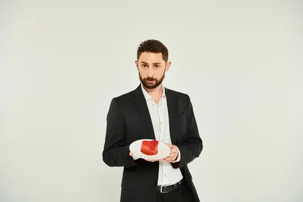 Elegante uomo barbuto che mostra piatto con torta a forma di sentire su grigio, delizioso trattamento di San Valentino — Foto stock