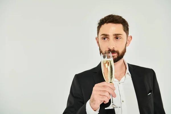 Homem barbudo elegante em terno preto bebendo champanhe e olhando para longe no dia dos namorados cinza, st — Fotografia de Stock