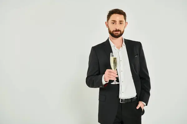 Uomo barbuto sorridente con bicchiere di champagne in piedi con la mano in tasca e guardando la fotocamera su grigio — Foto stock