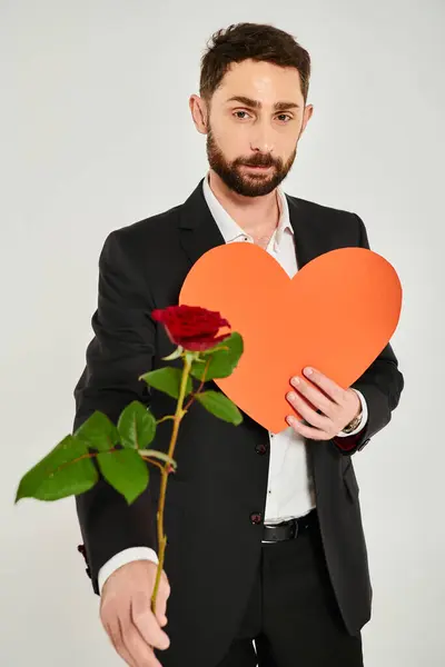 Стильний бородатий чоловік з червоною трояндою і помаранчевим паперовим серцем дивиться на камеру на сірому, Сен-Валентин — стокове фото