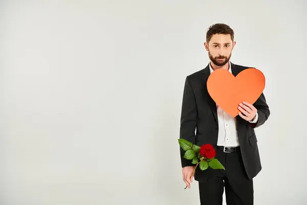 Eleganter bärtiger Mann mit roter Rose und riesigem Papierherz blickt am grauen Valentinstag in die Kamera — Stockfoto
