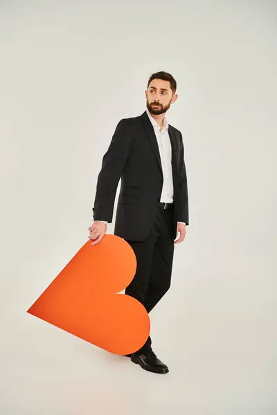 Пенсійний чоловік у чорному костюмі з величезним помаранчевим паперовим серцем, що дивиться на сірий, концепція священних валентин — стокове фото