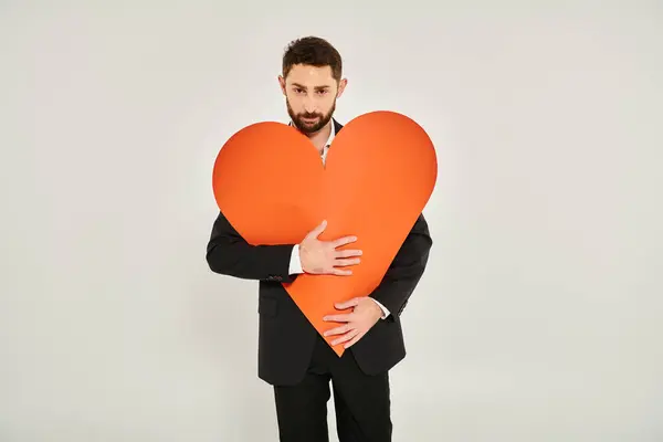 Bearded charismatic man embracing huge paper heart looking at camera on grey, Saint Valentines day — Stock Photo