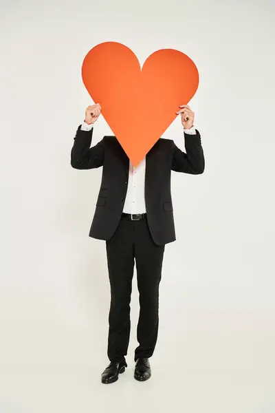 Elegant man in black suit obscuring face with huge orange paper heart on grey, st valentines concept — Stock Photo