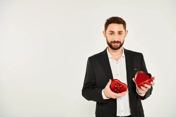 Uomo barbuto sorridente in abito nero apertura scatola regalo a forma di cuore rosso su grigio, st valentines presenti — Foto stock
