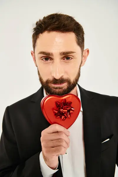 Щасливий бородатий чоловік показує подарункову коробку у формі серця з червоним бантом на сірому, день святого Валентина подарунок — стокове фото