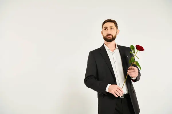 Мечтательный элегантный бизнесмен с красной свежей розой глядя в сторону на серый день Святого Валентина подарок — стоковое фото
