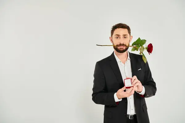 Bel homme avec rose rouge dans les dents montrant boîte à bijoux sur gris, Saint Valentin cadeau — Photo de stock