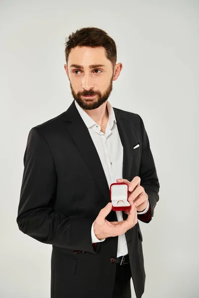 Позитивний бородатий чоловік показує ювелірну коробку з кільцем, дивлячись на сірий, присутній святий Валентин — стокове фото