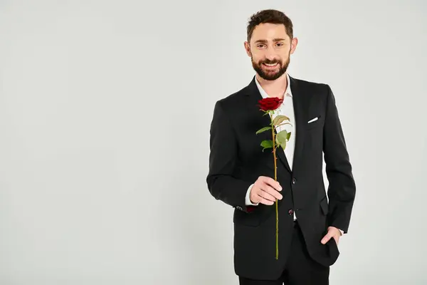 Allegro uomo barbuto in nero abbigliamento formale che tiene rosa rossa e guardando la fotocamera su sfondo grigio — Foto stock